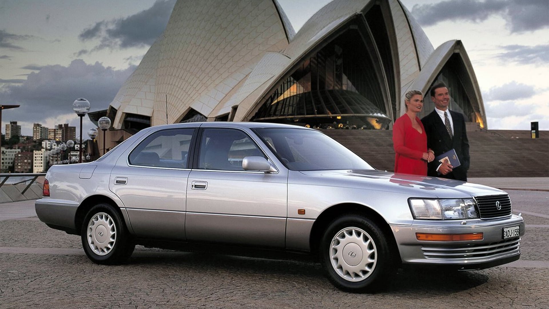 A man and woman stand with the first Lexus, silver, in front of the Sydney Opera House.
