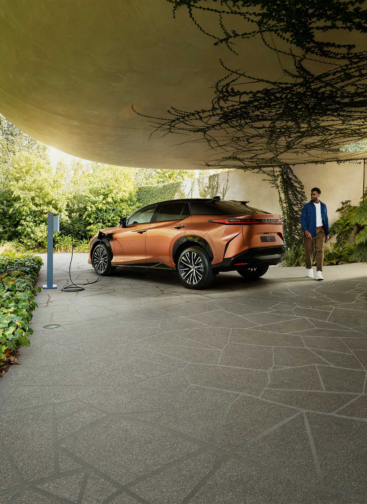 An copper Lexus RZ is parked and charging in a leafy green, vine-filled driveway. A man is seen walking around the rear of the car.